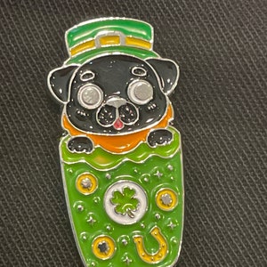 Puede incluir: Un pug negro con un sombrero de duende verde y amarillo asoma de una olla de oro verde. La olla está decorada con monedas de oro, una herradura y un trébol de cuatro hojas.