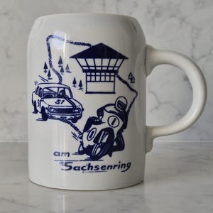 May include: White ceramic mug with a blue illustration of a race car, motorcycle, and a trackside structure. The text "am Sachsenring" is below the images. The mug has a curved handle.