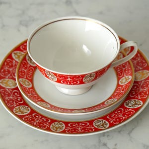 May include: A white teacup, saucer, and plate set with a red and gold decorative pattern. The teacup sits atop the saucer, which is on top of the plate. The set has a gold trim.
