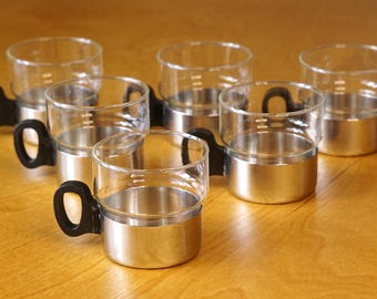 Op de afbeelding: Set van zeven heldere glazen bekers met roestvrijstalen bodems en zwarte handvatten. De bekers staan op een lichtbruin houten oppervlak. De bekers hebben maatstreepjes.