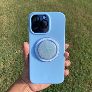 May include: A light blue phone case with a glittery silver pop-socket. The case has a camera cutout and a small emblem at the bottom. The pop-socket has a light purple border.
