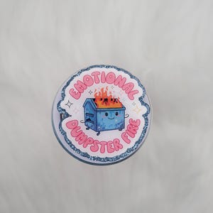 May include: A circular sticker with a blue glitter border featuring a cartoon blue dumpster on fire. The text EMOTIONAL DUMPSTER FIRE is written in pink bubble letters around the central illustration, accented by small white four-pointed star icons.