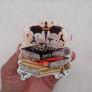 May include: A glittery sticker featuring three stacked books with the titles "Iron Flame", "Onyx Storm", and "Fourth Wing". A black dragon and other dragon silhouettes are above the books. The sticker has a sparkly, iridescent border.