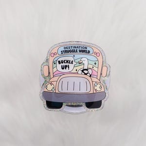 May include: A whimsical sticker featuring a cartoon bus with a goose driver. The bus is pink and gray with the text "DESTINATION STRUGGLE WORLD" and "BUCKLE UP!". The design includes a rainbow and stars.