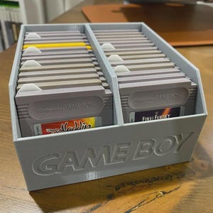 Può includere: Un supporto grigio per cartucce Game Boy pieno di più cartucce di gioco. Il supporto ha la scritta "GAME BOY" in rilievo sulla parte anteriore. Alcune cartucce sono etichettate "Aladdin" e "Final Fantasy".