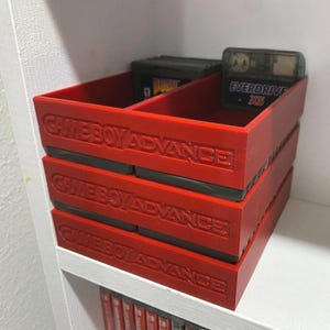 Puede incluir: Unidad de almacenamiento roja para cartuchos de Game Boy Advance. La unidad está apilada con tres compartimentos, cada uno con la etiqueta "GAMEBOY ADVANCE". El compartimento superior contiene cartuchos de juego, incluido uno con la etiqueta "EVERDRIVE".