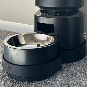May include: A black PETLIBRO automatic pet feeder with a stainless steel bowl. The feeder has a cylindrical design with a transparent food level indicator. The base includes a feeding bowl and a control panel. A power cord is visible.