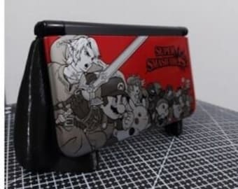 Peut inclure: Console Nintendo 3DS XL noire avec un couvercle à thème Super Smash Bros. rouge et blanc. Le couvercle présente une image de personnages de dessin animé du jeu.