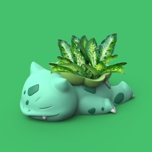 Puede incluir: Una maceta de cerámica verde con forma de personaje de dibujos animados durmiendo con una planta verde frondosa creciendo en su interior.