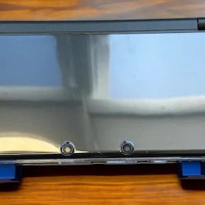 K&ouml;nnte beinhalten: Eine blaue Nintendo 3DS XL Handheld-Spielekonsole mit einer transparenten Schutzh&uuml;lle. Die Konsole ist geschlossen und der obere Bildschirm ist sichtbar.