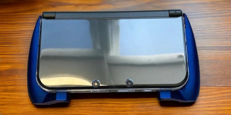 K&ouml;nnte beinhalten: Eine blaue Nintendo 3DS XL Handheld-Spielekonsole mit einer transparenten Schutzh&uuml;lle. Die Konsole ist geschlossen und der obere Bildschirm ist sichtbar.