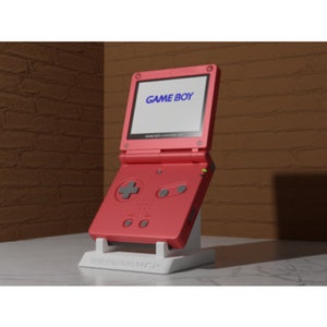 Könnte beinhalten: Eine rote Game Boy Color Handheld-Videospielkonsole mit einem weißen Ständer. Die Konsole hat einen schwarzen Bildschirm mit dem Text "GAME BOY" darauf.