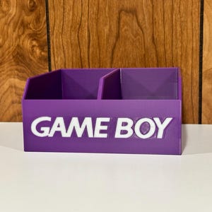 以下が含まれることがあります： 前面に白い「GAME BOY」の文字が入った、3つのコンパートメントを備えた紫色のオーガナイザー。 オーガナイザーはしっかりとした素材でできており、小さなアイテムを収納できるように設計されています。 背景は木製の表面です。
