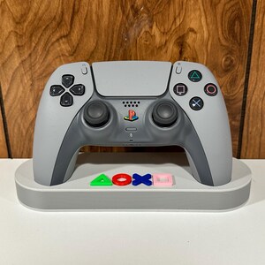 Op de afbeelding: Een witte PlayStation 5 DualSense-controller met zwarte knoppen en een PlayStation-logo op het touchpad, rustend op een witte oplaadstandaard met het PlayStation-logo in kleurrijke letters.