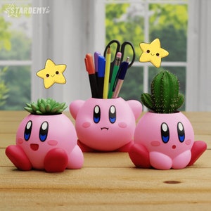 May include: Three pink planter pots shaped like the character Kirby from the video game series Kirby. The pots are holding small plants, including a cactus and two succulents. The pots have a cute, cartoonish design with big eyes and a small mouth.