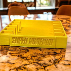 May include: A yellow plastic storage case with slots for video game cartridges. The case has the words "SUPER NINTENDO" printed on the front.