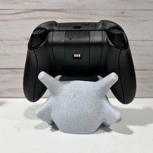 May include: A black Xbox controller is sitting on a white, speckled, 3D printed stand. The controller has the Xbox logo on the back.