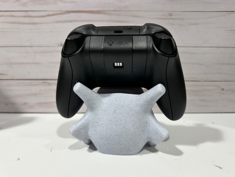 May include: A black Xbox controller is sitting on a white, speckled, 3D printed stand. The controller has the Xbox logo on the back.