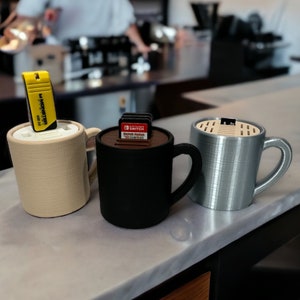 May include: Three ceramic mugs with different colored glazes. The mugs are designed to hold small items like game cartridges and USB drives. The mugs are sitting on a light gray countertop.