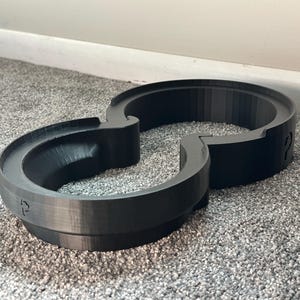Pode incluir: Uma pista de brinquedo preta impressa em 3D, que se assemelha a um oito. A pista tem sec&ccedil;&otilde;es curvas e uma borda elevada. A pista &eacute; feita de um material preto mate e assenta num tapete cinzento.