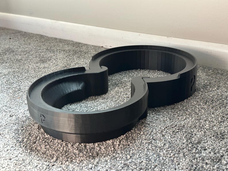 Pode incluir: Uma pista de brinquedo preta impressa em 3D, que se assemelha a um oito. A pista tem sec&ccedil;&otilde;es curvas e uma borda elevada. A pista &eacute; feita de um material preto mate e assenta num tapete cinzento.