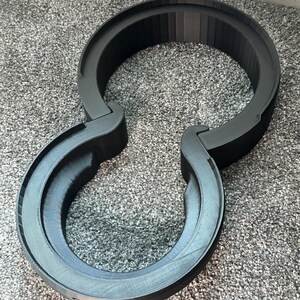 Pode incluir: Um suporte de fone de ouvido preto impresso em 3D. O suporte tem um design circular com dois bra&ccedil;os curvos que se estendem dos lados. O suporte &eacute; feito de um material preto fosco e foi projetado para segurar fones de ouvido.