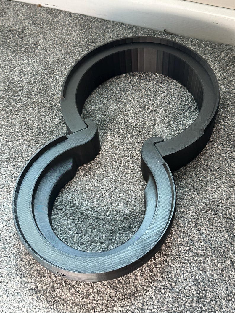 Pode incluir: Um suporte de fone de ouvido preto impresso em 3D. O suporte tem um design circular com dois bra&ccedil;os curvos que se estendem dos lados. O suporte &eacute; feito de um material preto fosco e foi projetado para segurar fones de ouvido.