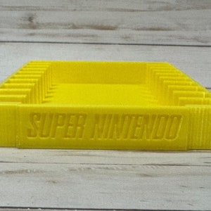 May include: A yellow plastic storage case for Super Nintendo game cartridges. The case has slots for multiple cartridges and the words "SUPER NINTENDO" are printed on the front.
