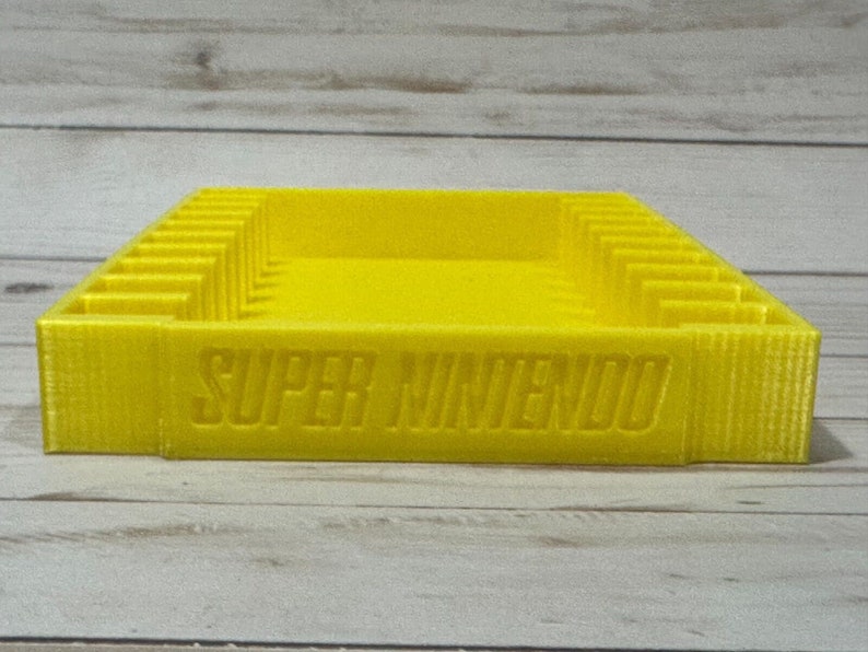 May include: A yellow plastic storage case for Super Nintendo game cartridges. The case has slots for multiple cartridges and the words "SUPER NINTENDO" are printed on the front.
