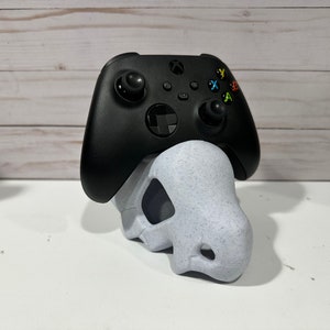 Pode incluir: Um controlador Xbox preto está sentado em um suporte branco em forma de caveira. O suporte é feito de um material salpicado.