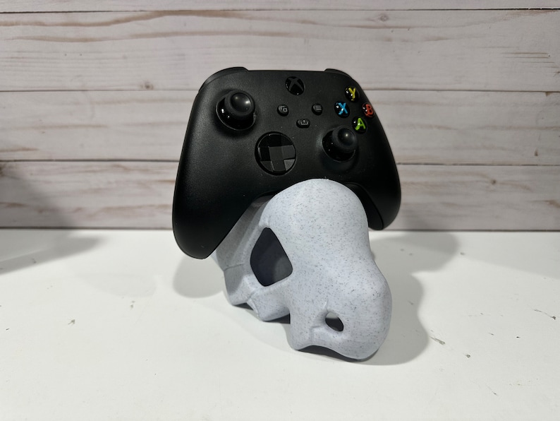 May include: A black Xbox controller is sitting on a white skull-shaped stand. The stand is made of a speckled material.