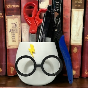May include: White ceramic Harry Potter themed pencil holder with a lightning bolt and glasses. The holder is filled with a pair of scissors, a pen, and a blue tool.
