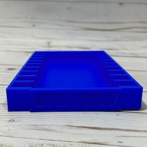 May include: A blue plastic storage case for Super Nintendo game cartridges. The case has 12 slots for cartridges and the words "SUPER NINTENDO" are embossed on the front.
