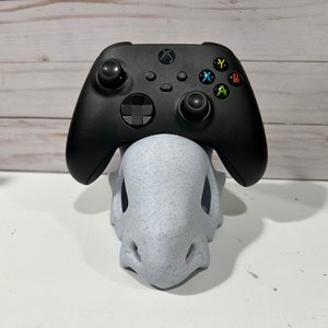 May include: A black Xbox controller sits on a white, speckled, 3D printed stand. The controller has a black and gray color scheme with colored buttons.