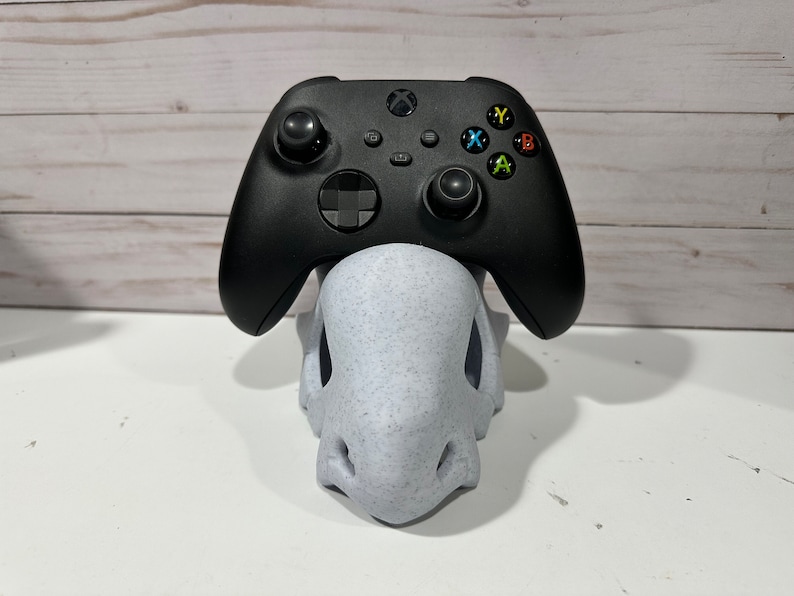 May include: A black Xbox controller sits on a white, speckled, 3D printed stand. The controller has a black and gray color scheme with colored buttons.