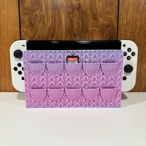 May include: A purple and pink 3D printed game cartridge holder with 12 slots for Nintendo Switch game cartridges. The holder is designed to look like a knitted pattern.