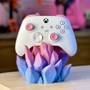 May include: A white Xbox controller with pink buttons sits on a stand made of blue and pink crystals.