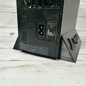 May include: Black PlayStation 3 console with a black stand. The console has a power button, a USB port, and an Ethernet port. The stand has the text "De Proxima" printed on it.