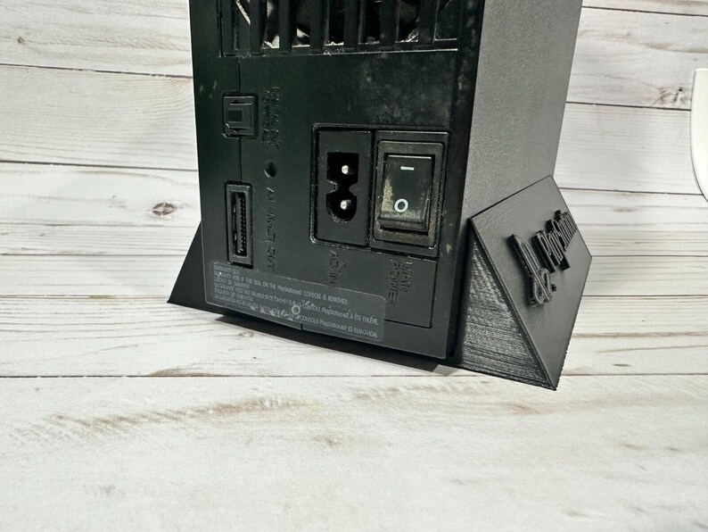 May include: Black PlayStation 3 console with a black stand. The console has a power button, a USB port, and an Ethernet port. The stand has the text "De Proxima" printed on it.