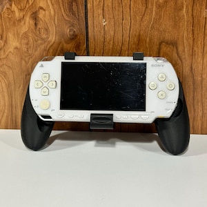 以下が含まれることがあります： 黒いグリップと黒い画面を備えた白いソニーPSPハンドヘルドゲーム機。コンソールには、方向パッド、アクションボタン、電源ボタンがあります。コンソールは黒いスタンドに取り付けられています。
