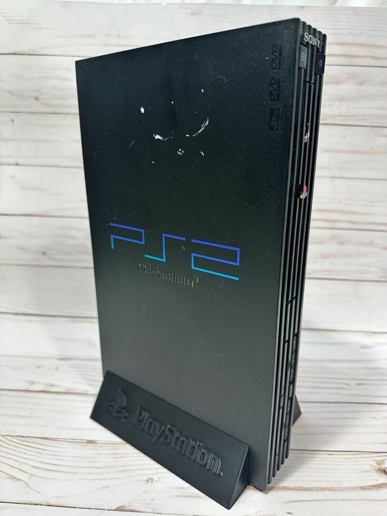 May include: A black Sony PlayStation 2 console with a blue and white PlayStation 2 logo. The console is standing on a black stand with the PlayStation logo.