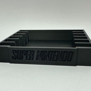 May include: A black plastic storage case with slots for video game cartridges. The case has the words "SUPER NINTENDO" printed on the front.