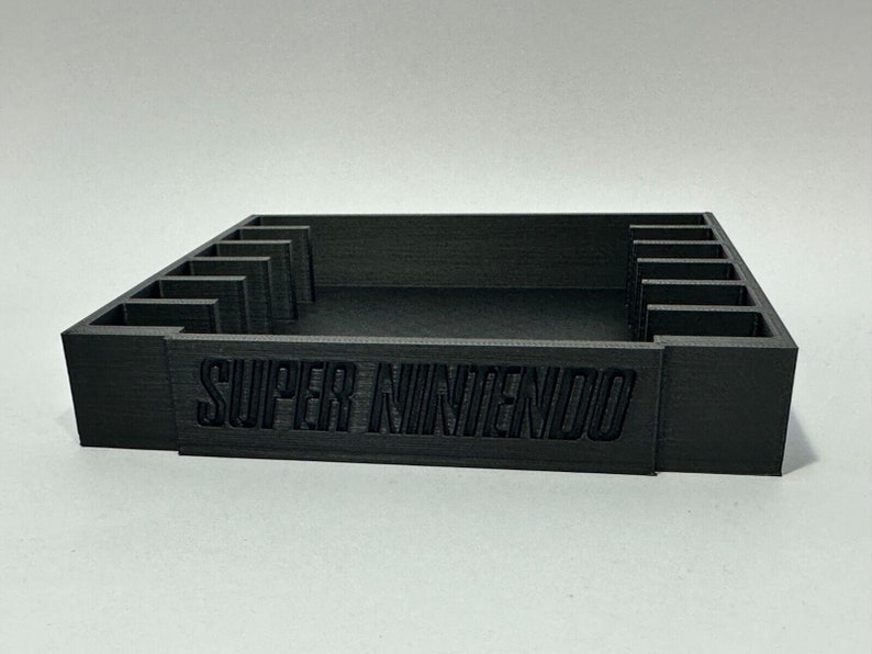 May include: A black plastic storage case with slots for video game cartridges. The case has the words "SUPER NINTENDO" printed on the front.