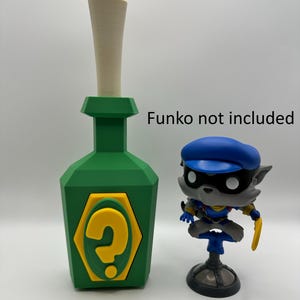 Puede incluir: Una botella verde con un signo de interrogación amarillo en la parte delantera. La botella tiene una tapa blanca y está hecha de plástico. La botella está al lado de una figurita gris y azul de un lobo que lleva un sombrero azul. El texto "Funko not included" es visible en la imagen.