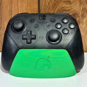May include: A black Nintendo Switch controller with a green Animal Crossing themed base. The controller has a D-pad, two analog sticks, and four face buttons. The base has a leaf design.