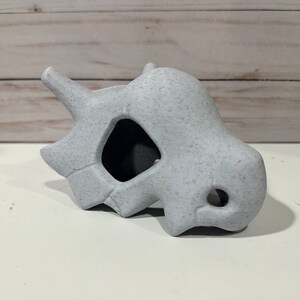 May include: A white ceramic skull with a textured surface. The skull has a large hole in the front and a smaller hole on the side.