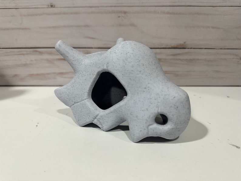May include: A white ceramic skull with a textured surface. The skull has a large hole in the front and a smaller hole on the side.