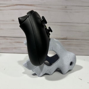 May include: A black video game controller is resting in a white plastic stand. The stand is shaped like a dinosaur head.
