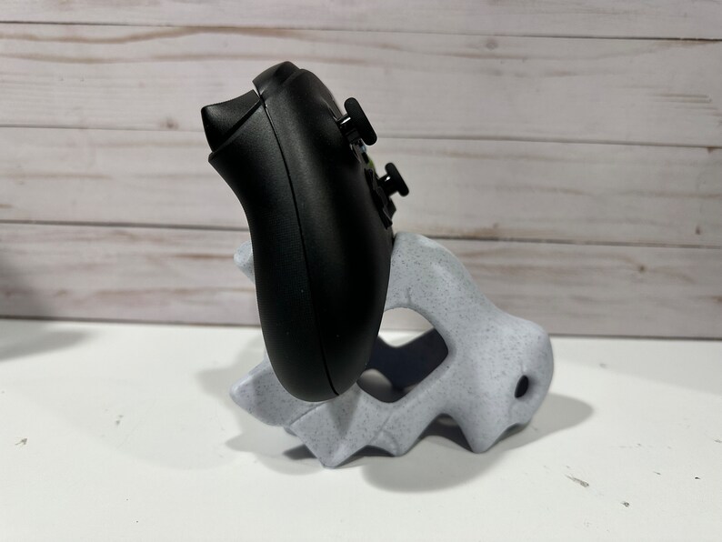 May include: A black video game controller is resting in a white plastic stand. The stand is shaped like a dinosaur head.