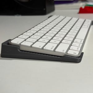 May include: A white keyboard with a grey base is angled upwards. The keyboard has white keys with black lettering. The base is a dark grey colour. The keyboard is on a white surface.